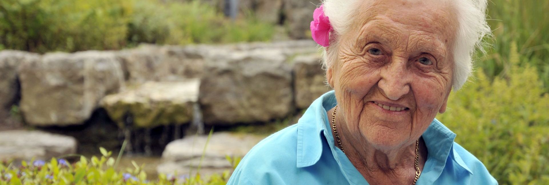 Ältere Frau mit rosa Blüte im Haar lächelnd im Garten